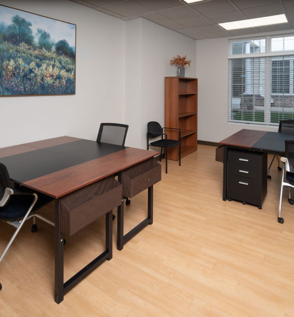 Private office featuring a desk with chair facing a window, framed landscape artwork, bookshelf, and additional seating with light wood flooring.