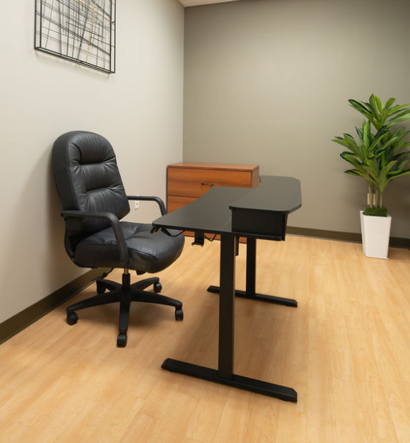 Private office with black executive chair, height-adjustable desk, wooden credenza, and potted plant on light hardwood flooring.