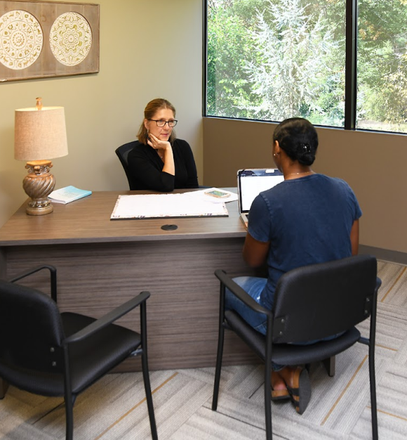 Small private meeting office with a woman and client seated at a desk, laptop open, framed wall art, and large window with garden view.